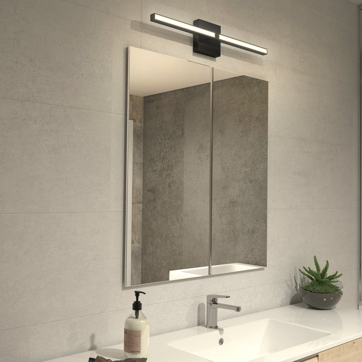 A modern bathroom with light gray tile walls features a large rectangular mirror and the Artika Convertible Pro 24-in Integrated LED Vanity Light Bar above it, plus a sink with chrome faucet, soap dispenser, a potted succulent, and a wooden counter.