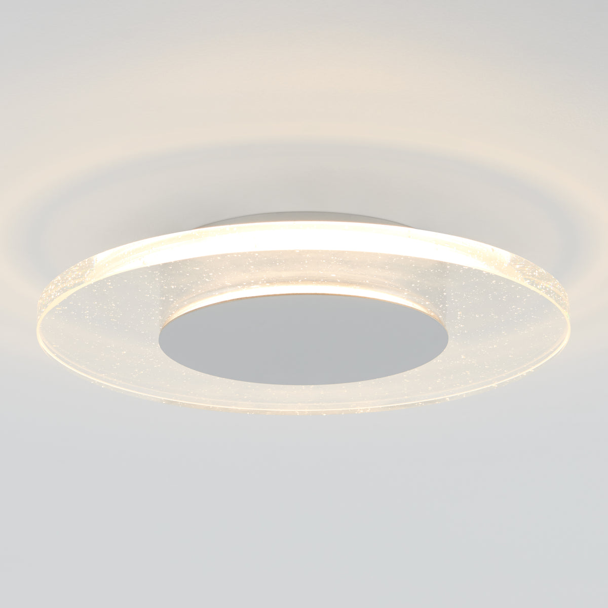 The Artika Essence Disk LED flush mount is a modern circular ceiling fixture featuring a glowing ring and metallic center, providing soft light and subtle reflections on the white ceiling.