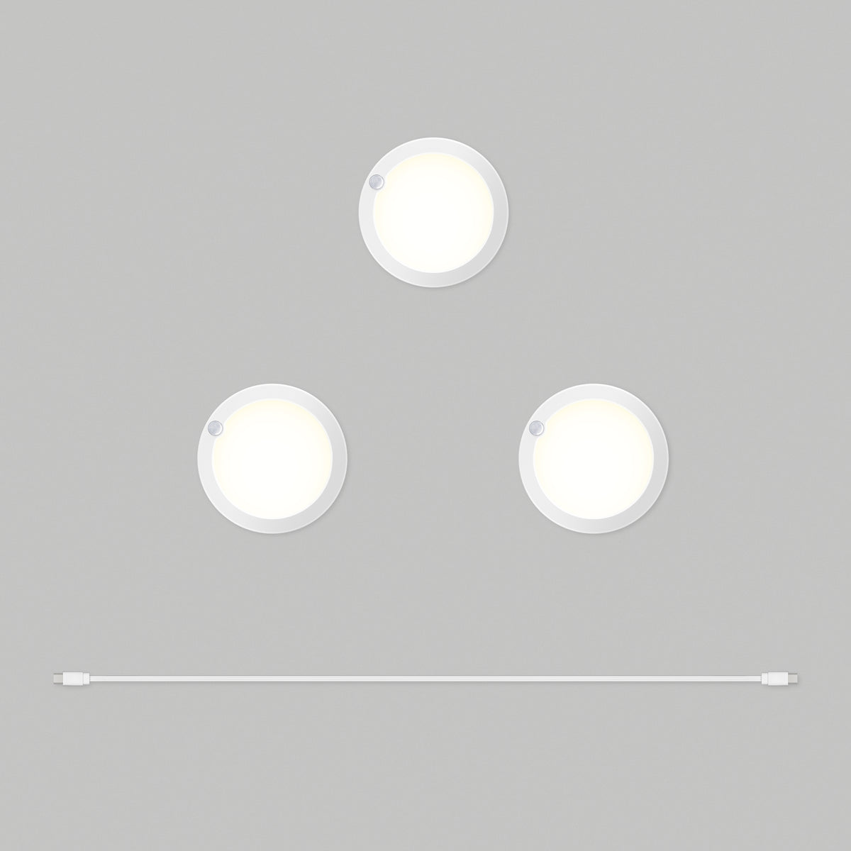Three round white Artika Accent under cabinet LED light 3CCT puck lights are arranged in a triangle on a gray background, with a white USB cable positioned horizontally below them.