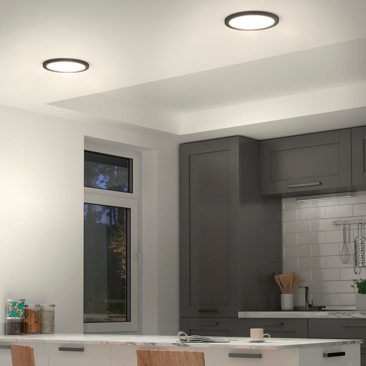 A modern kitchen features Artikas Europa Disk 9 in. LED flush mount lights, gray cabinets, a window, white subway tile backsplash, and a white countertop with a mug and plate. Wooden chairs are partially visible in the foreground.