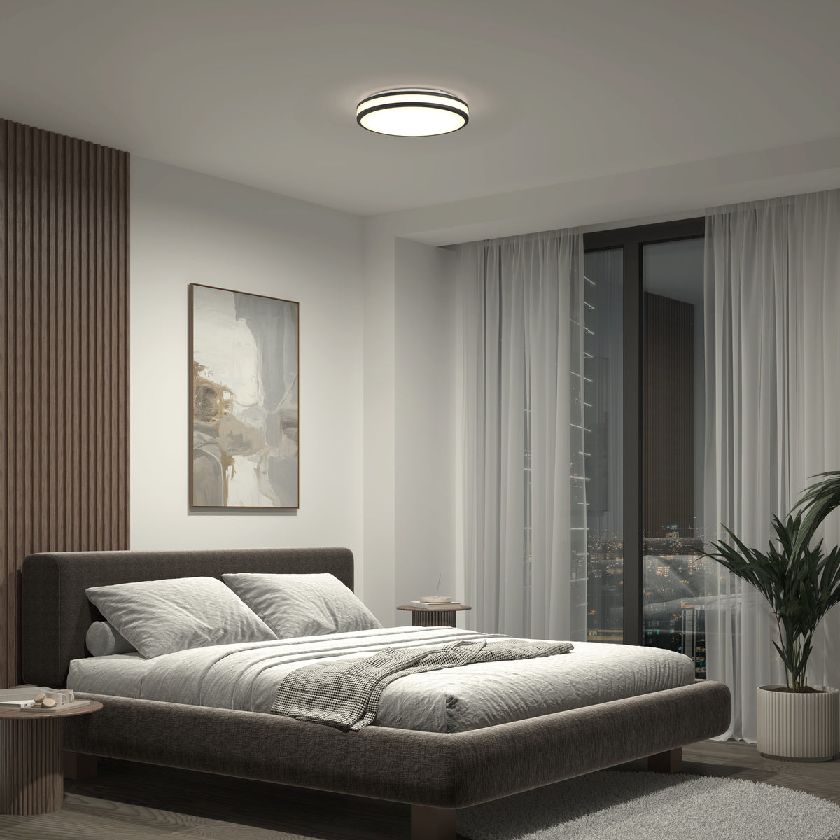 A modern bedroom with a low, dark bed, gray bedding, abstract wall art, nightstands, a potted plant, floor-to-ceiling windows revealing a nighttime city view, and an Artika Hemisphere 15 in. LED flush mount light with 5 CCT adjustable brightness on the ceiling.