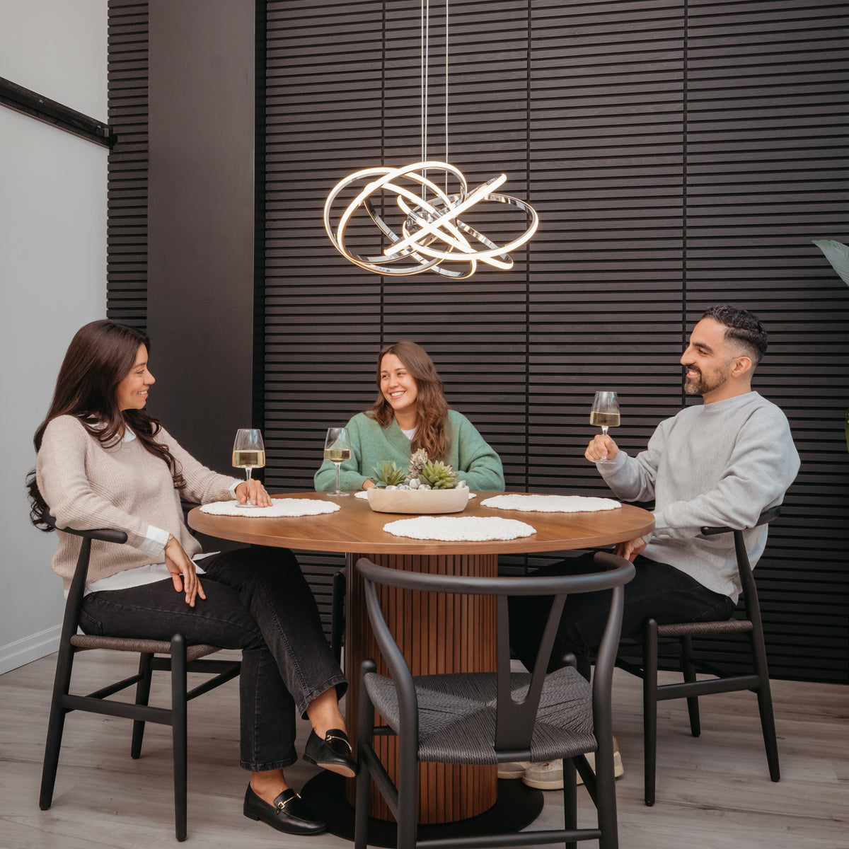 Three people sit around a round wooden table, smiling with wine glasses. Above them hangs the Artika Maelstrom modern LED pendant light 5 CCT, illuminating the space against a dark, textured wall.