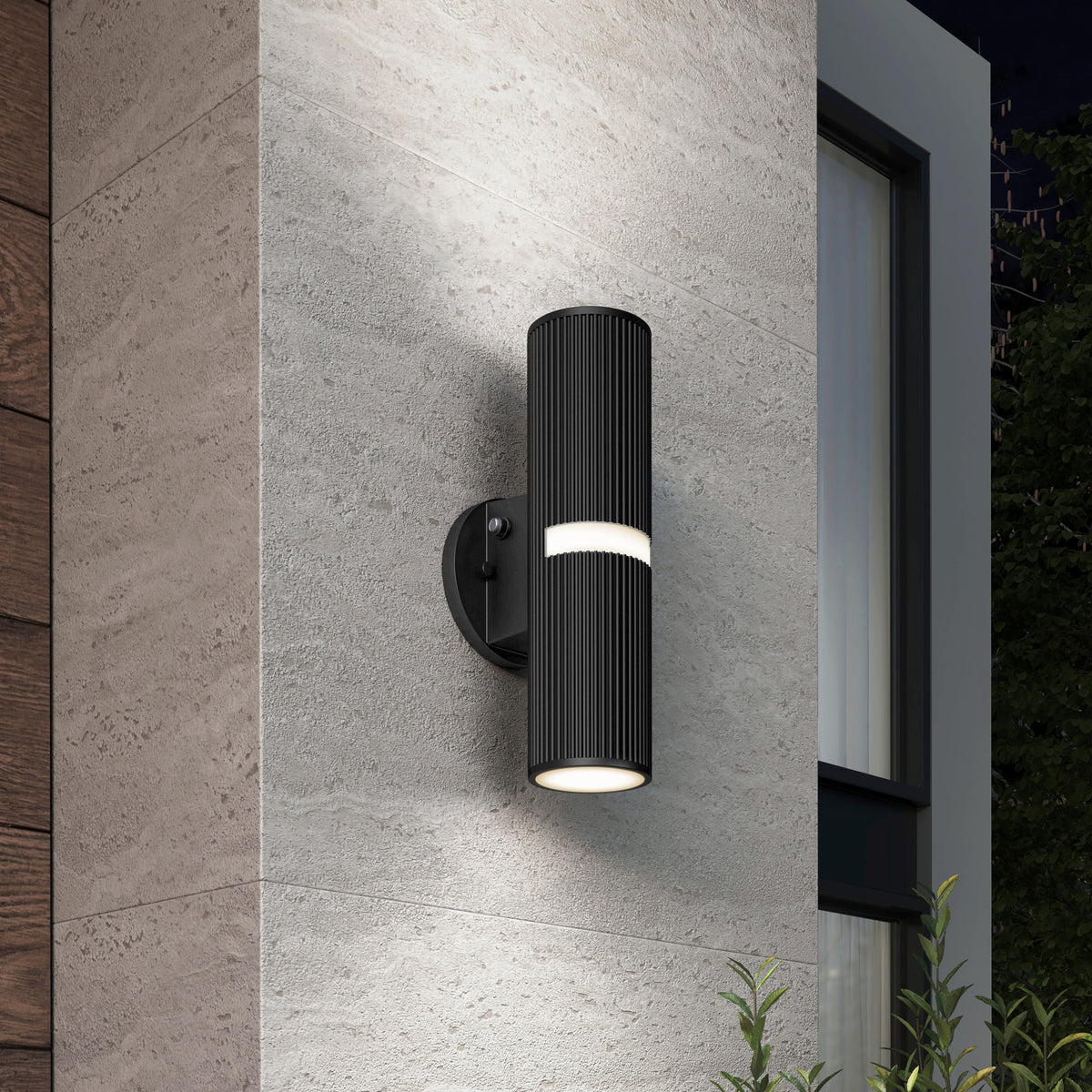 The Artika Groove LED indoor-outdoor wall light, a modern black cylindrical fixture with ribbed texture, is mounted on a light gray stone wall near a window, casting light up and down. Some plants appear at the bottom right.