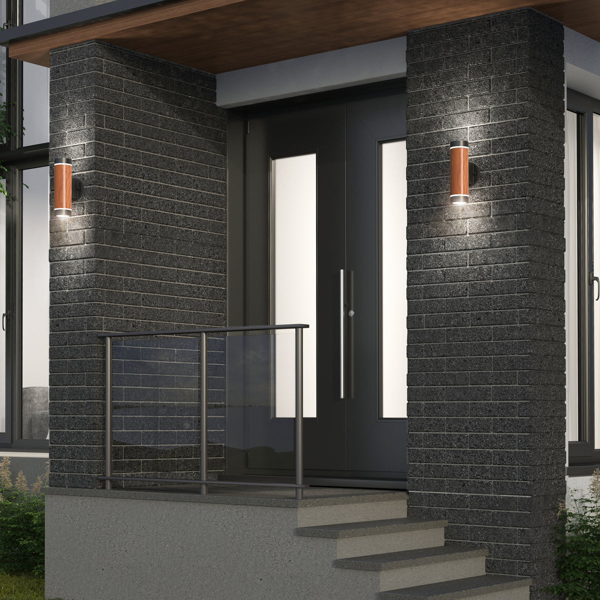 Modern house entrance with black brick walls, double glass front doors, metal railing, and two Artika C7 indoor-outdoor LED wall lights (3 CCT) illuminating the entryway. Steps lead up to the small porch area.