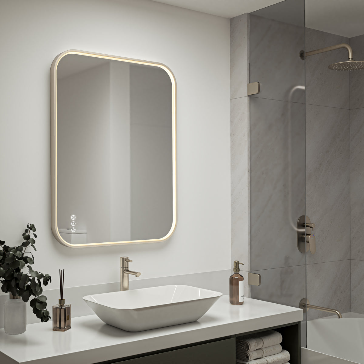 Modern bathroom featuring the Artika Royce Dimmable LED Mirror (3 CCT, horizontal/vertical mount) above a white sink with gold faucet, soap dispenser, potted plant, and reed diffuser. Gold-accented glass shower appears in the background.