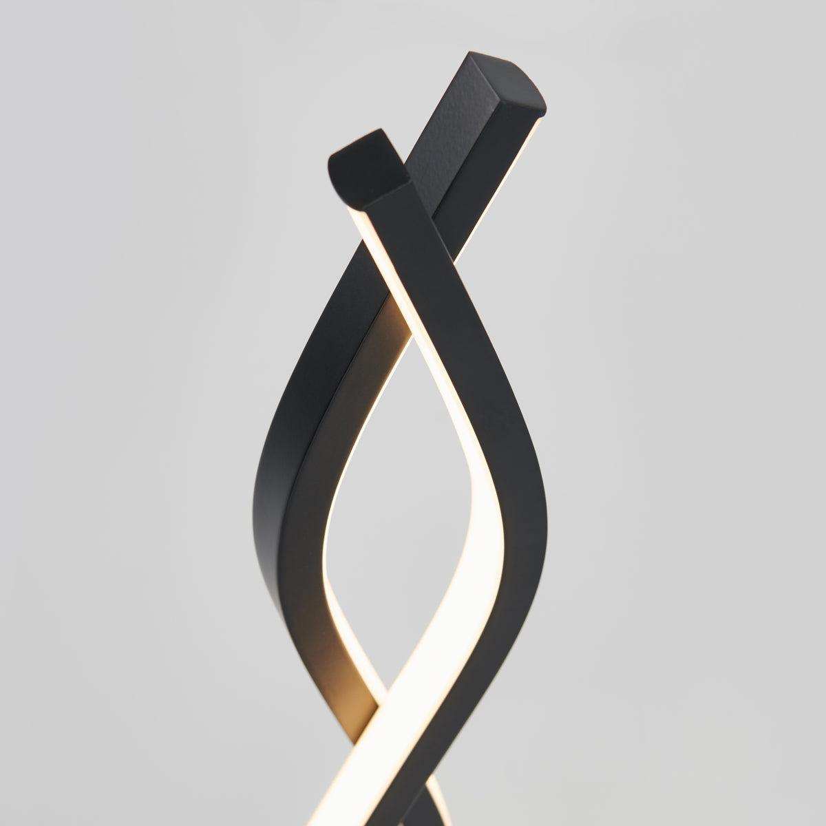 Artikas Wave LED Table Lamp features two modern black curved fixtures that elegantly twist together, emitting a soft glow against a plain gray background.