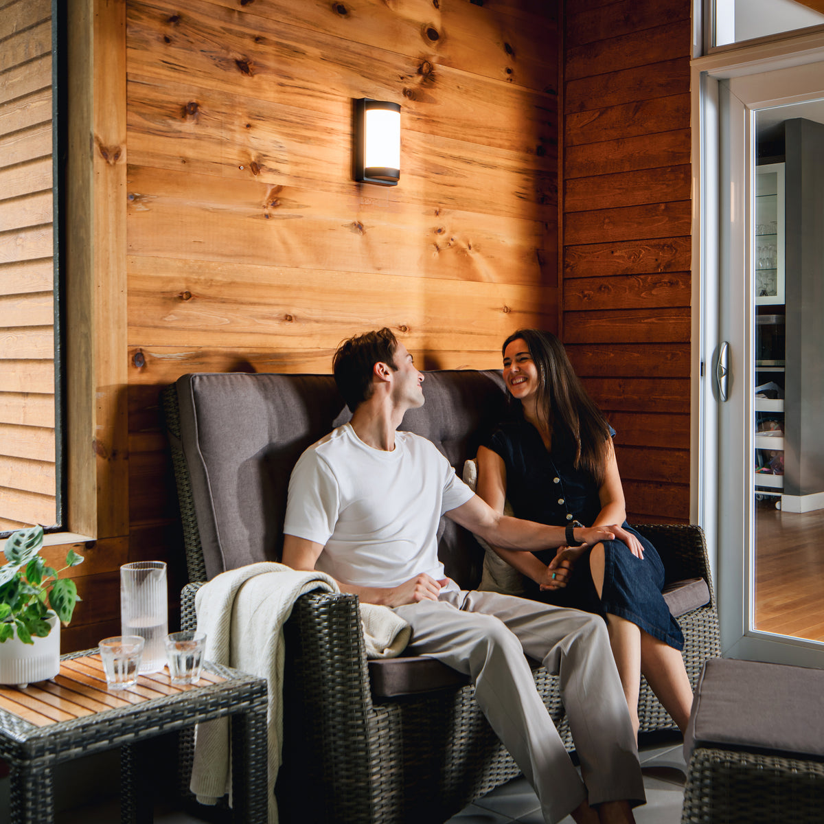 A couple relaxes on a patio sofa, enjoying drinks under Artika’s Fogo LED wall light in a cozy room with plants.