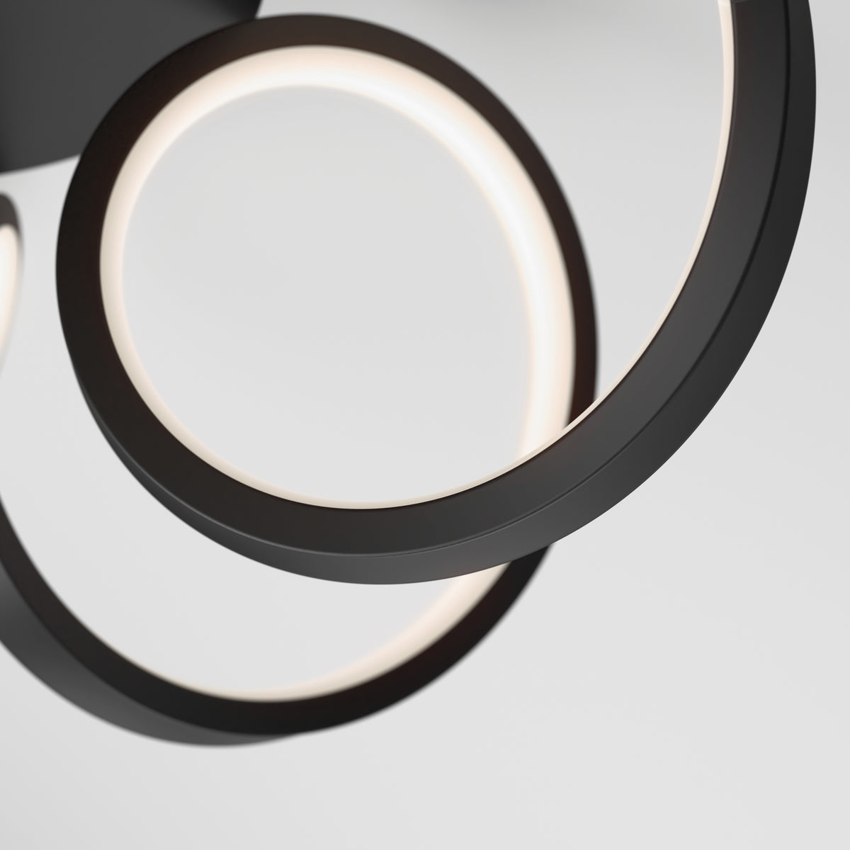 A close-up of the Artika Swirl Ribbon LED pendant light showcases two intertwined black metal rings with integrated LEDs, casting a soft white glow against a plain light background.