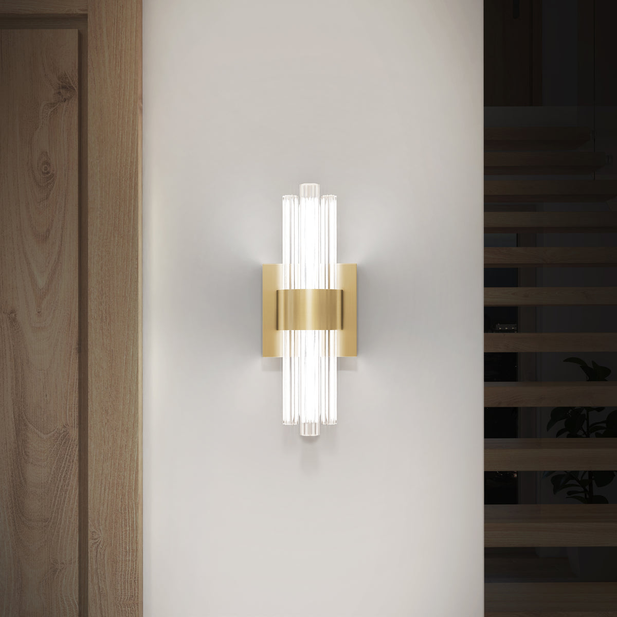 The Artika Dorado contemporary LED wall sconce 3 CCT features vertical glass rods and a gold metallic center, mounted on a white wall by a wooden door and stairs. This luxury fixture provides soft, diffused illumination.