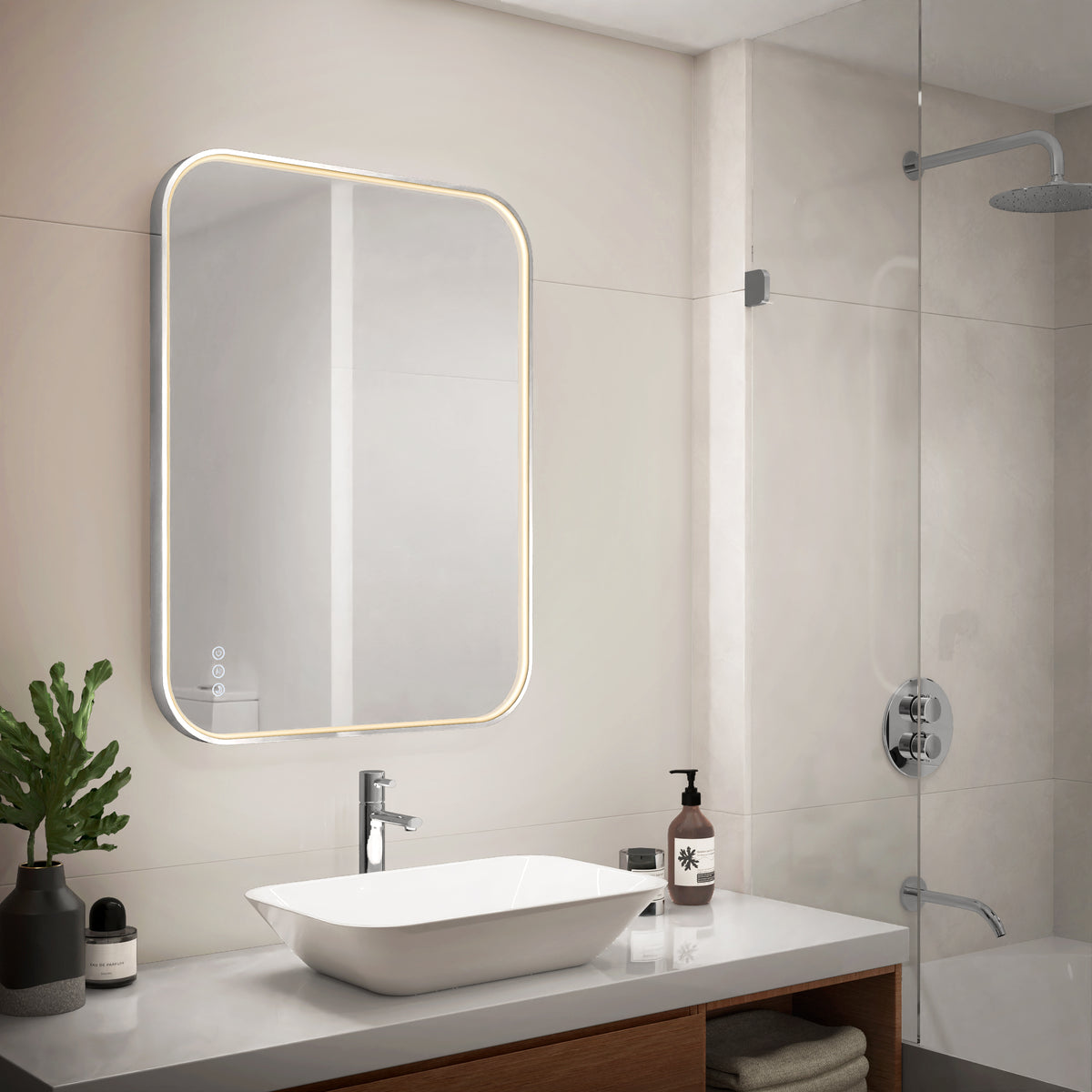 Modern bathroom with the Artika Royce Dimmable LED Mirror (3 CCT, horizontal/vertical mount) above a white sink, plant, and soap dispensers. To the right is a glass shower enclosure with a rainfall showerhead.