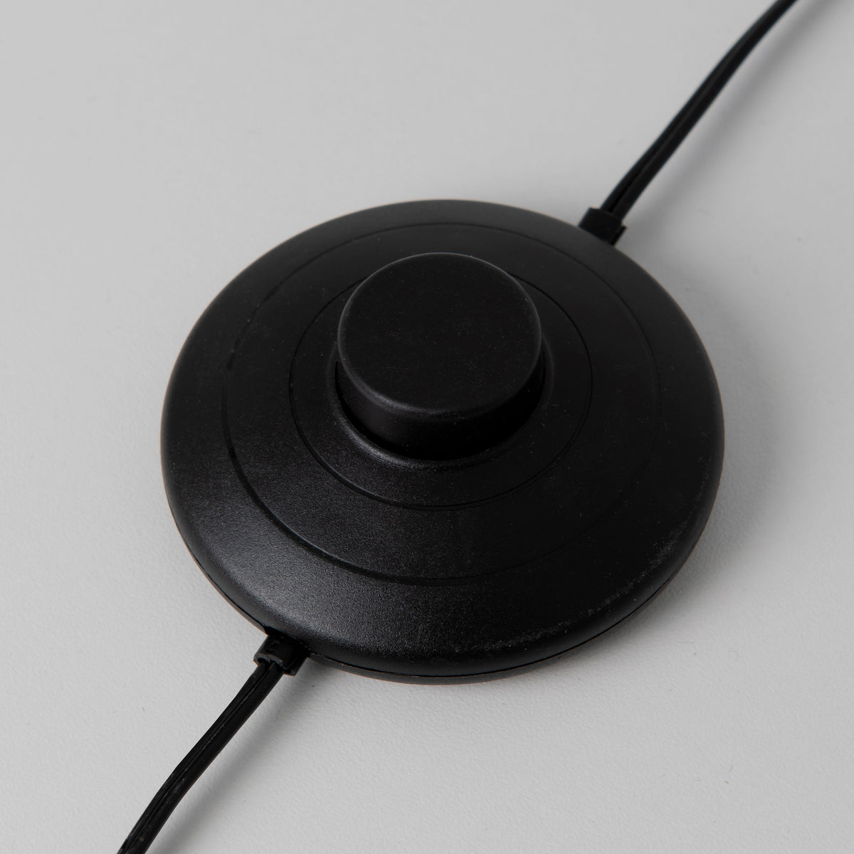 A black circular foot switch with a central button, connected to two black wires, is shown on a light gray surface for the Swirl modern LED floor lamp by Artika.