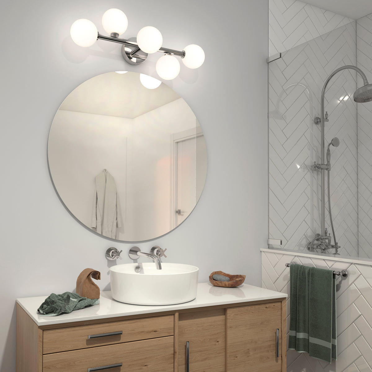 Modern bathroom with a round mirror above a wooden vanity, white vessel sink, and silver faucet. The Artika Bloom 27-in 3-Light Modern LED Vanity Light adds style above, complemented by a green towel and chevron-tiled glass shower.