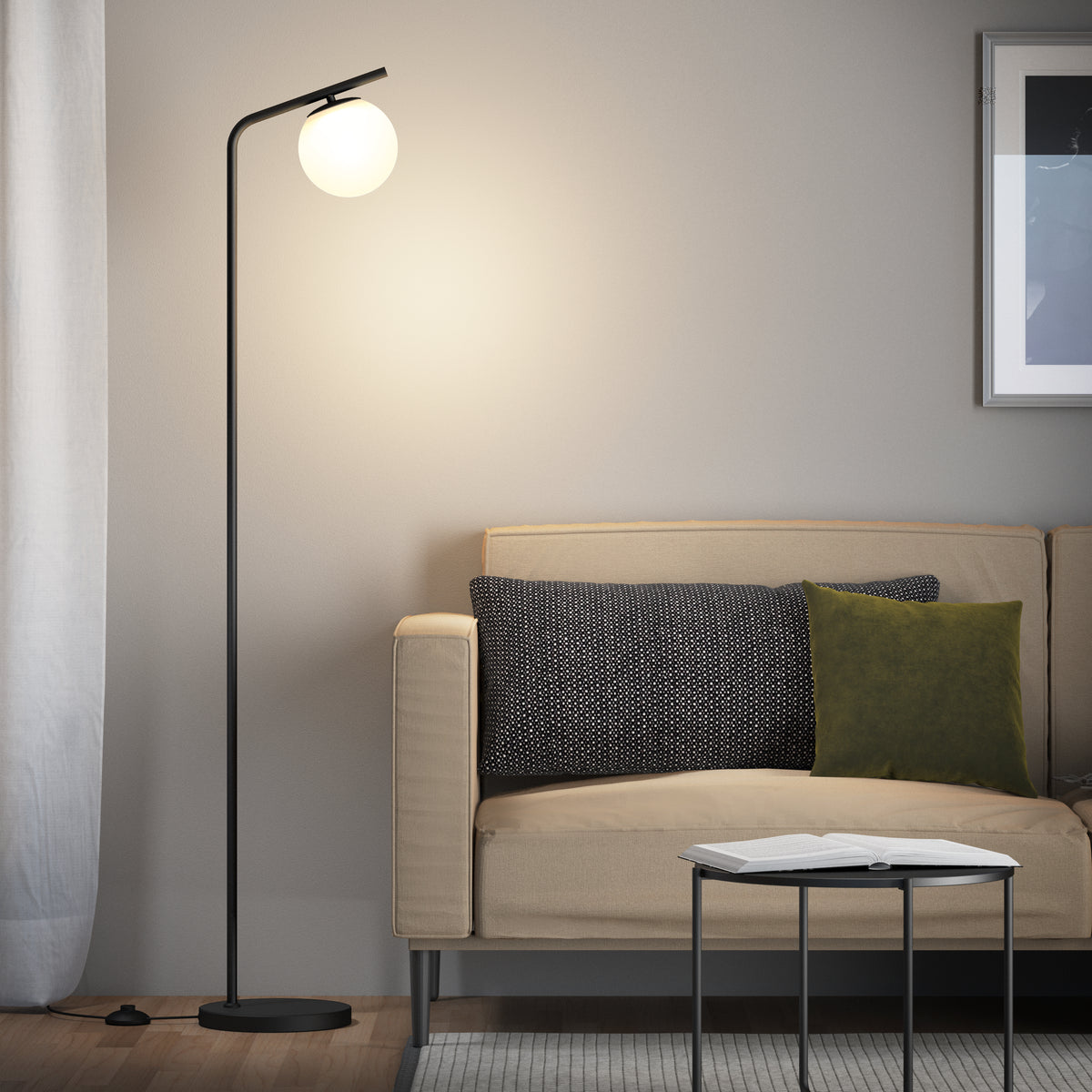 A modern living room with a beige sofa, dark and green throw pillows, a round black side table holding an open book, and the Artika Clayton dimmable LED modern mid-century floor lamp with a glowing round white shade beside the sofa.