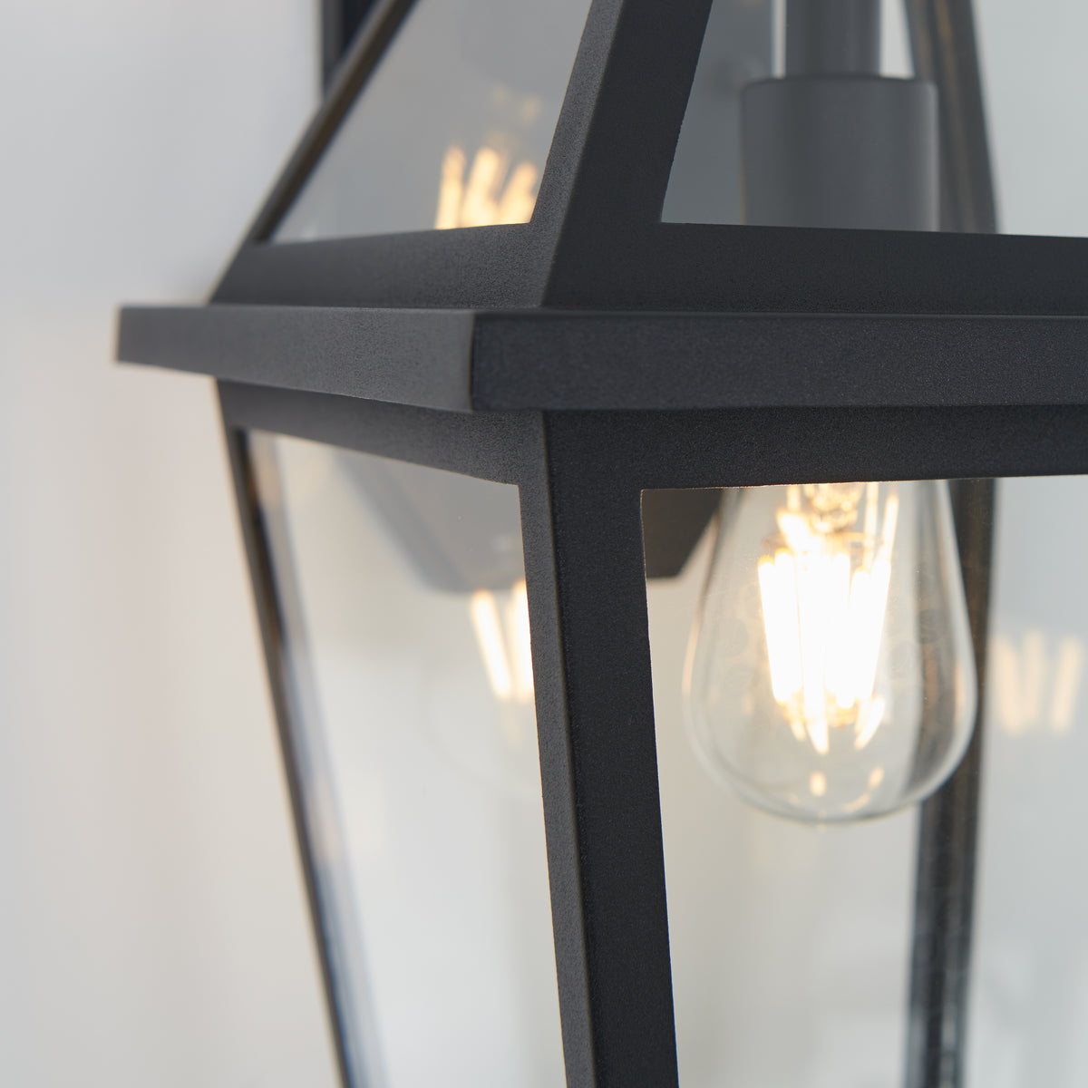 Close-up of the Artika Colonial outdoor lantern wall light, featuring black metal and glass with a vintage-style exposed filament bulb, mounted on a light-colored wall.