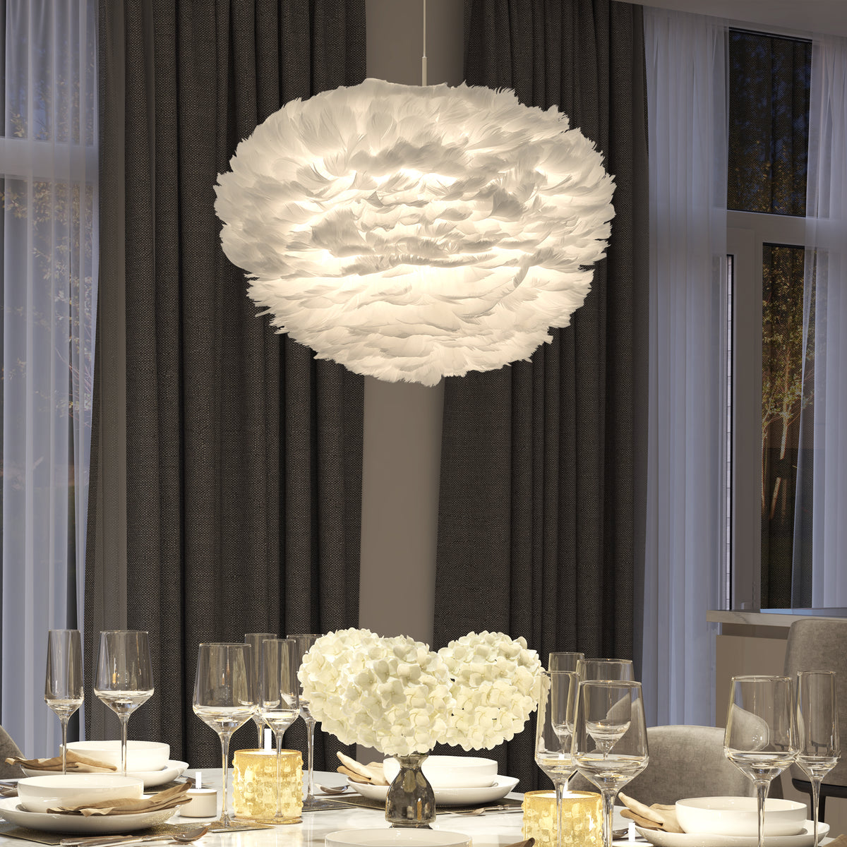 A modern dining table set with glassware and plates sits beneath the Artika Plume LED pendant light. The scene is accented by two white flower arrangements, dark curtains, and large windows.