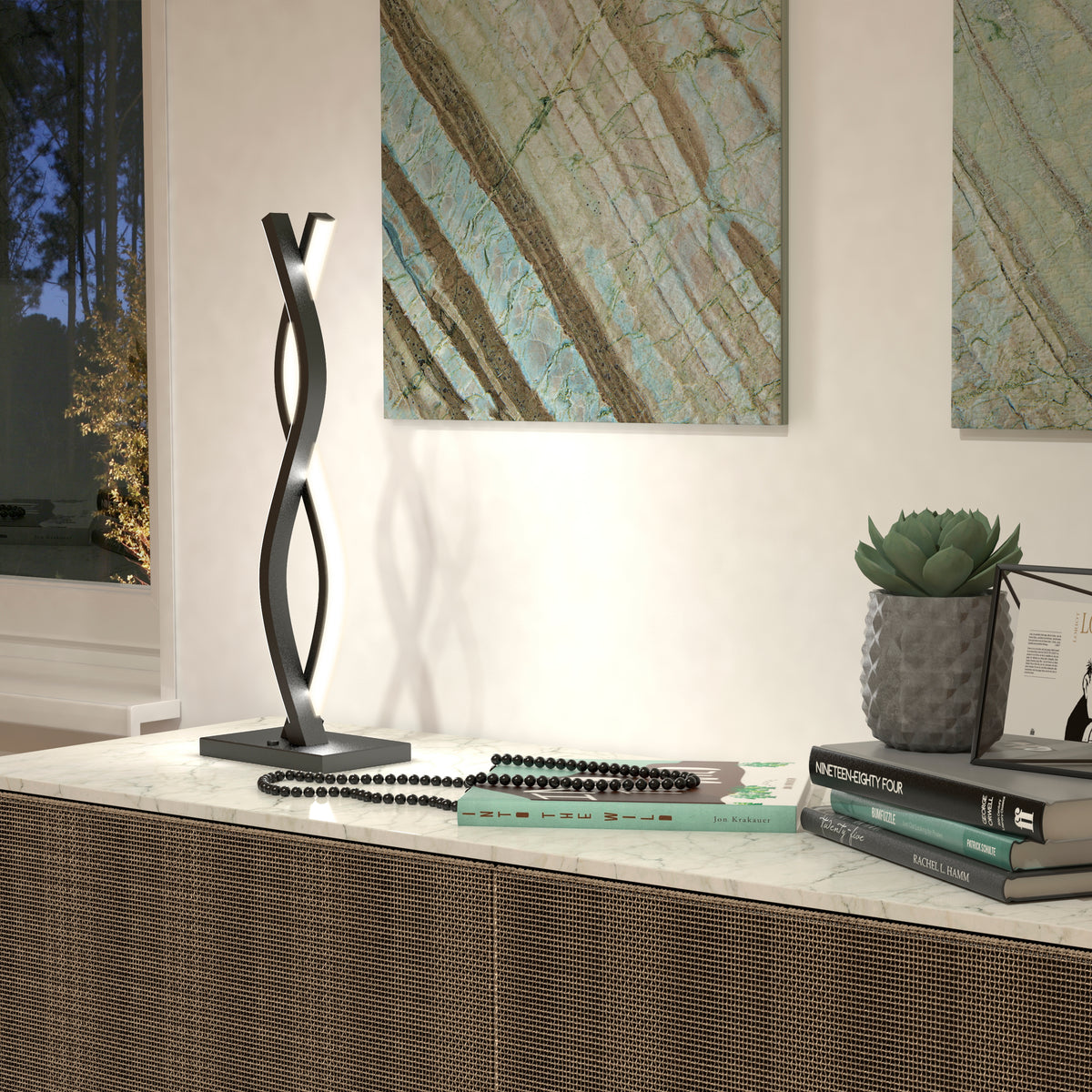 A modern table features the Artika Wave LED Table Lamp with twisted black metal, a stack of books, a beaded necklace, a succulent in a textured pot, and two abstract art pieces on a neutral wall in the background.