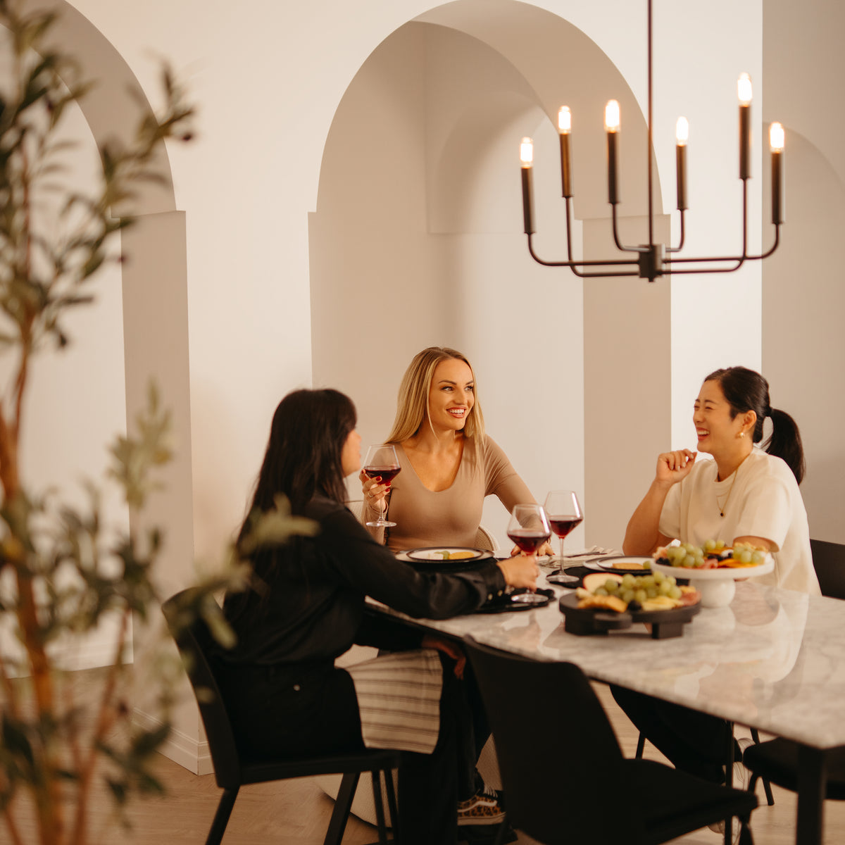 Three women enjoy red wine and conversation at a dining table set with food, while the Artika Manchester 6-light pendant casts a warm glow overhead in a stylish room with arched openings.