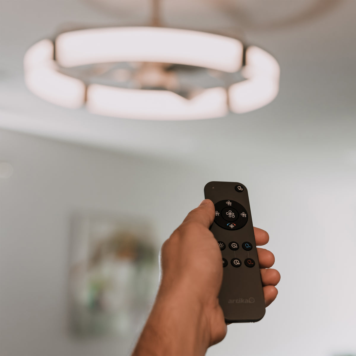 A hand holds a remote for the Artika Sunnyvale 28 in. LED modern ceiling fan, shown in a softly lit room.