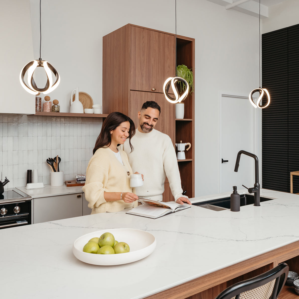 A woman and man smile together in a modern kitchen with wood cabinets and a bowl of green apples, illuminated by Artikas Vivaldi LED pendant light 3 CCT hanging above the island.