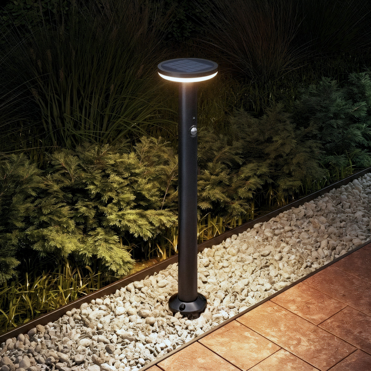 Artika’s Urbia Solar LED Bollard with Motion Sensor lights up at night beside white rocks, plants, and a tiled walkway.