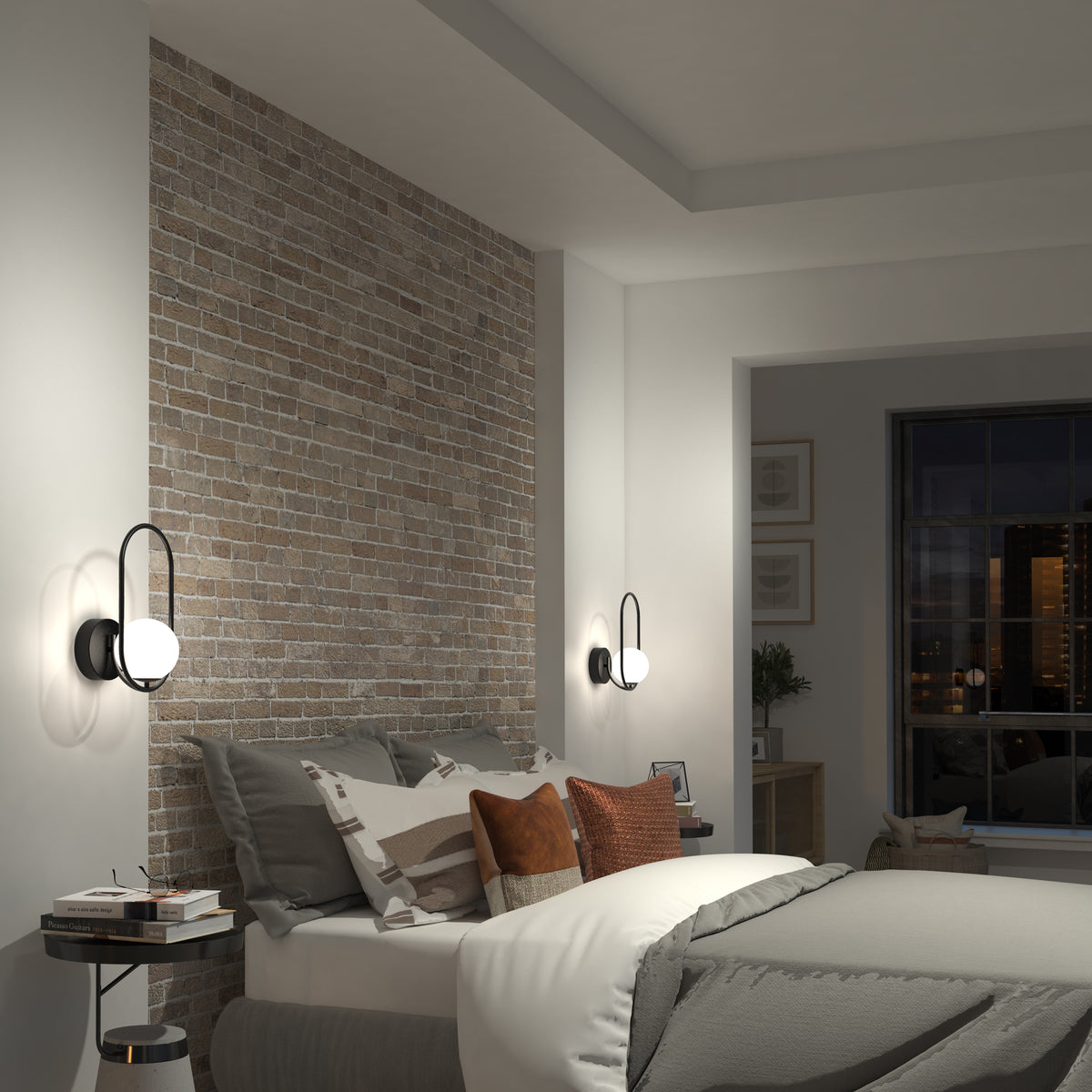 A modern bedroom with a brick accent wall, a neatly made bed with gray and brown pillows, Artika’s Bloom LED Sconce 3 CCT, a round side table, and a window revealing city buildings at night.