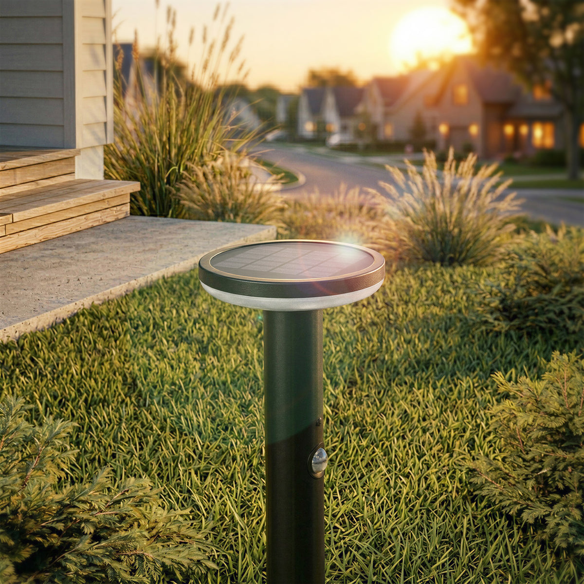 An Artika Urbia Solar LED Bollard with Motion Sensor stands in grass by a porch, houses and sunset visible behind.
