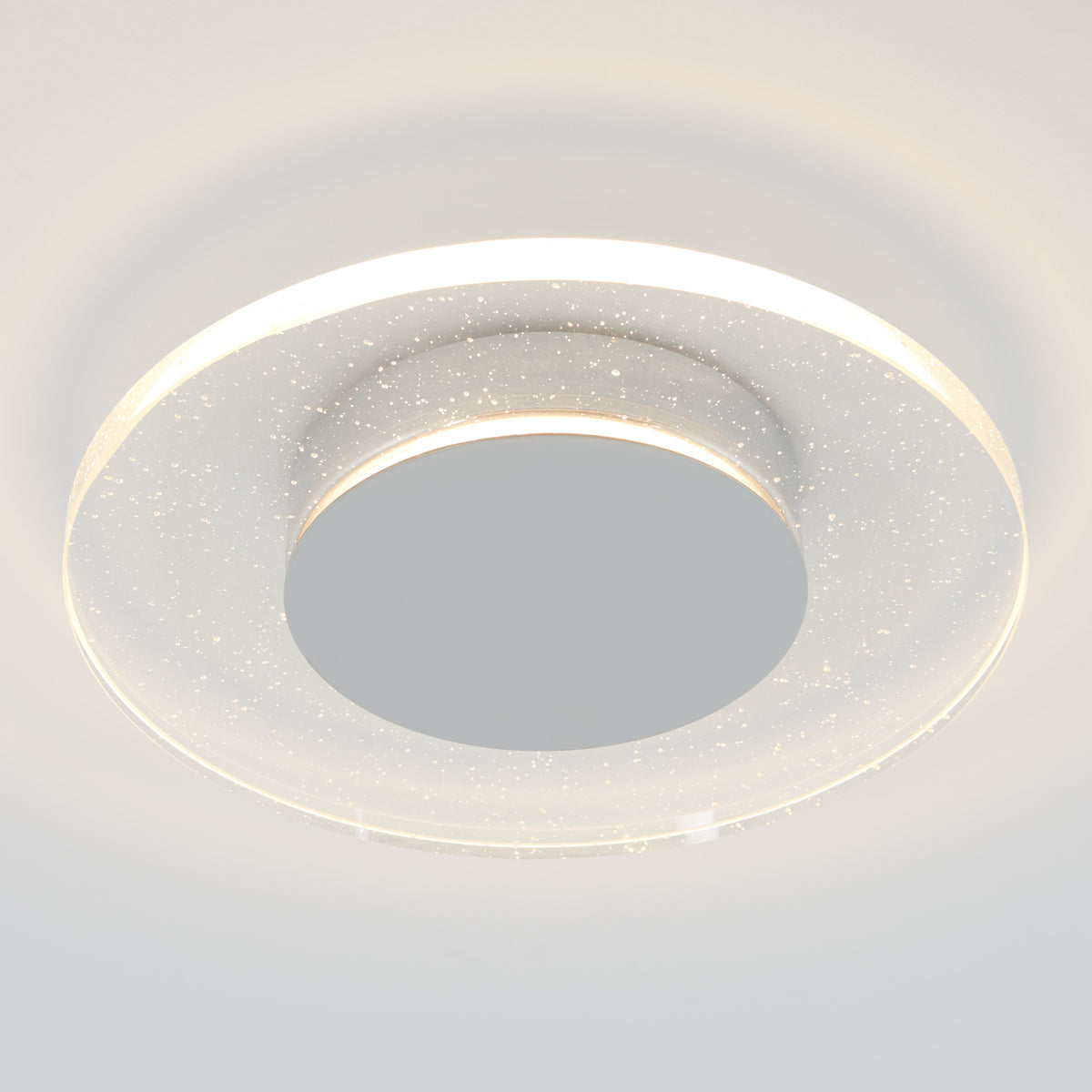 The Artika Essence Disk LED flush mount features a frosted, semi-transparent ring with tiny star-like dots, casting a soft white glow on pale ceilings for a modern look.