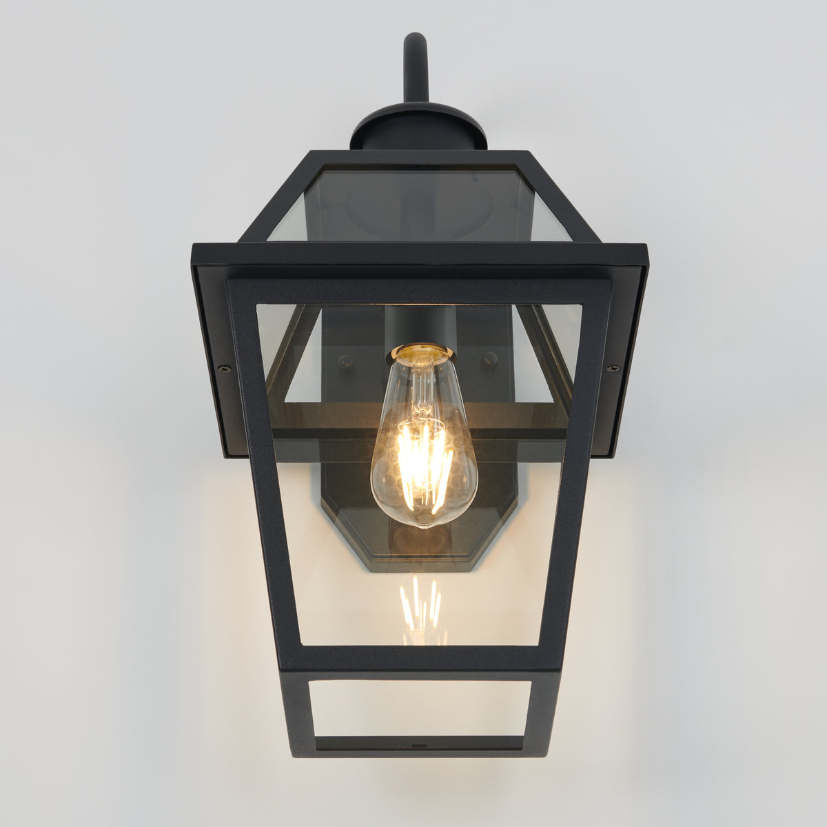 The Artika Colonial outdoor lantern wall light is mounted on a white wall, showcasing a visible glowing filament bulb inside its rectangular black metal and glass enclosure.