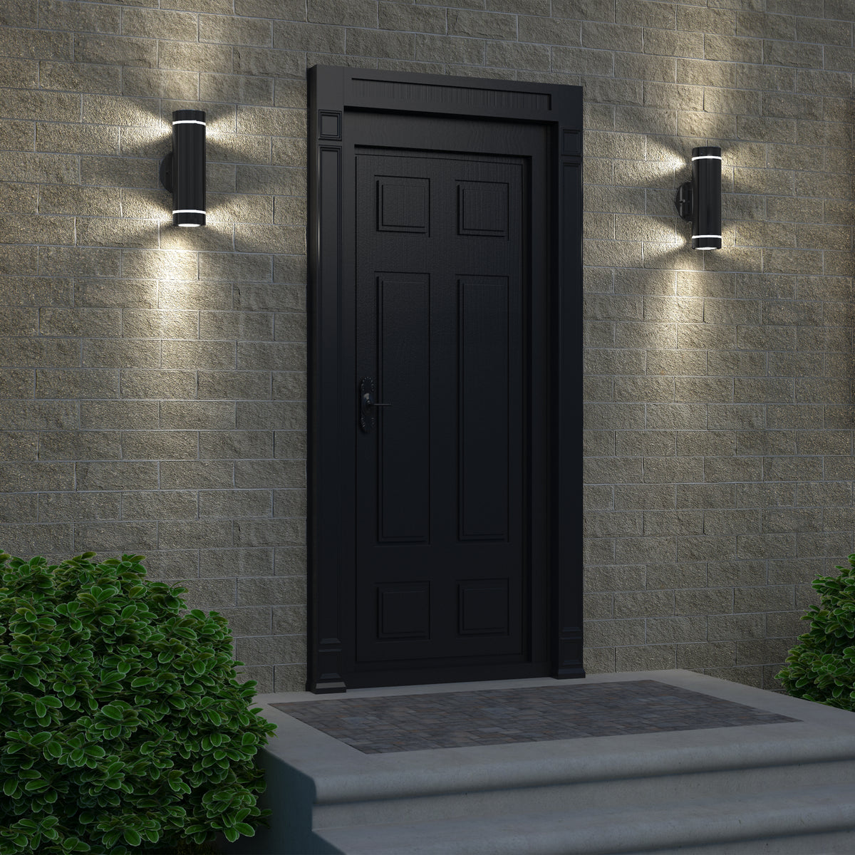 A black door with decorative panels sits in a light brick wall, illuminated on each side by Artika’s C7 indoor-outdoor wall lights. Concrete steps lead up to the entrance, flanked by green bushes.