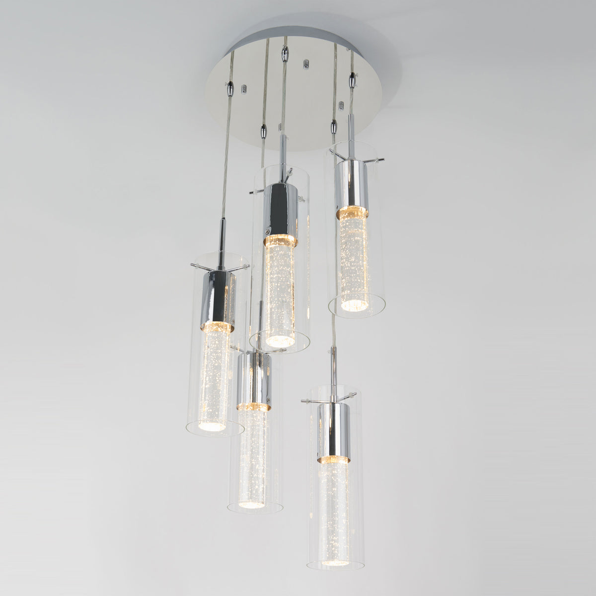 The Artika Bubble Swirl 5-light pendant features five cylindrical glass shades with sparkling details, suspended at different heights from a round white ceiling mount against a plain, light-colored background.