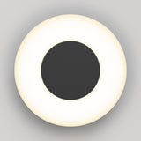 A circular wall light with a black center and a glowing white ring around it, mounted on a light gray background, creating a minimalist and modern appearance.