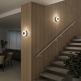 A modern staircase with gray steps beside a wood-paneled wall, featuring two circular wall lights. In the background, a dining area with a table and chairs is visible, and a potted plant sits at the bottom of the stairs.