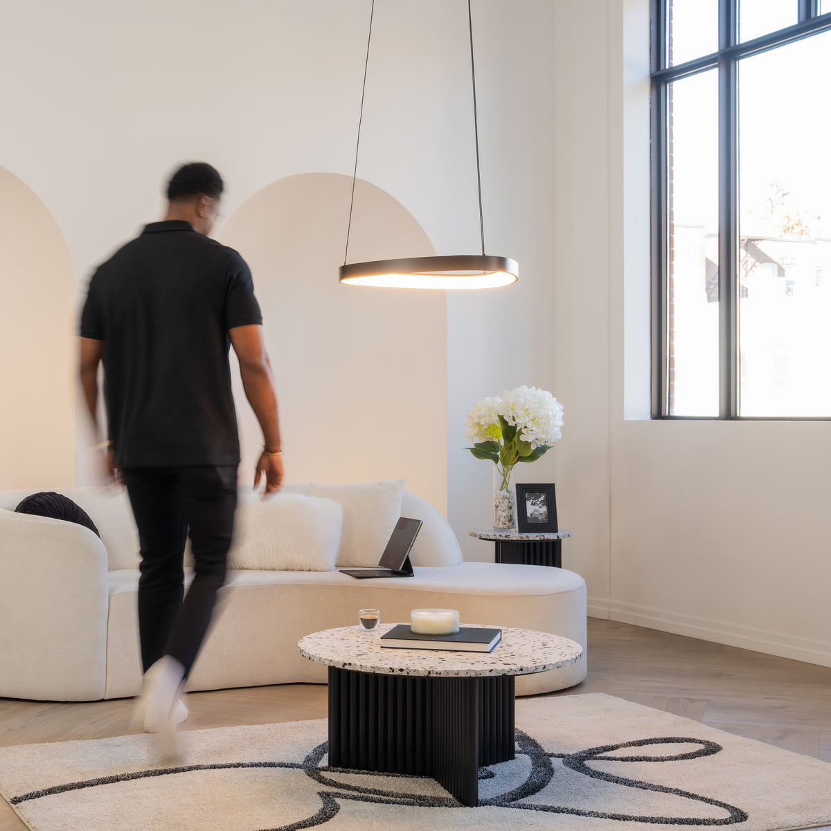 A man in black clothes walks through a modern living room with a white sofa, round coffee table, side table with flowers, large window, and a hanging circular light fixture.