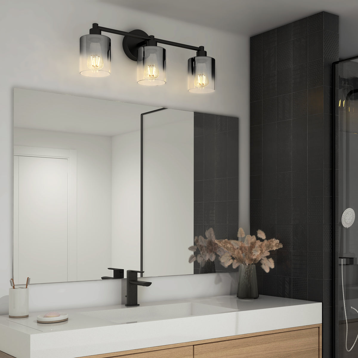 A modern bathroom with a large mirror, wooden vanity, black faucet, and a three-light fixture with glass shades above the mirror. A vase with dried plants sits on the white countertop. Dark tiles cover one wall.