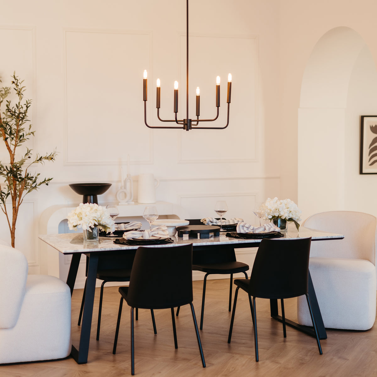 A modern dining room features a rectangular table set for four with black chairs, white floral centerpieces, neutral armchairs, a black chandelier, and minimalist decor in a bright, elegant space.