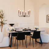 A modern dining room features a rectangular table set for four with black chairs, white floral centerpieces, neutral armchairs, a black chandelier, and minimalist decor in a bright, elegant space.