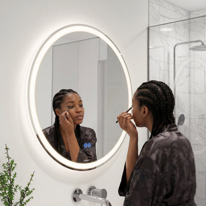 A woman with braided hair applies eyeliner while using the Artika Ayla 28 in. LED mirror 3 CCT with defogger in a modern bathroom with a glass shower, small potted plant, and adjustable lighting.