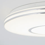 Close-up of a modern, round ceiling light fixture with a white and silver design, glowing softly against a white ceiling. The brand name artika is faintly visible on the edge.