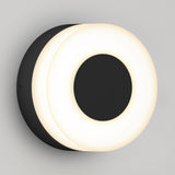 A modern wall sconce with a circular black base and a glowing white ring around a solid black center, mounted on a light gray wall.