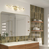 Modern bathroom with a wood vanity, gold faucet, and large rectangular mirror. Wall features greenish tiles, floating shelves with towels and decor, and a sculptural gold light fixture above the mirror.