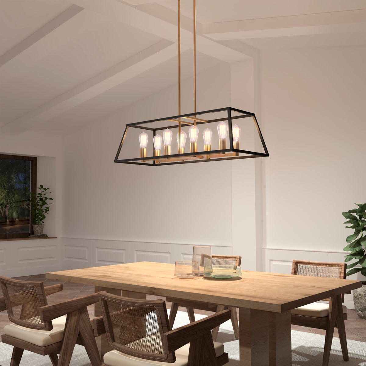 A modern dining room with a rectangular wooden table, four woven chairs, and a black and gold geometric chandelier with exposed bulbs hanging above. The room features white walls, plants, and a window.