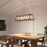 A modern dining room with a rectangular wooden table, four woven chairs, and a black and gold geometric chandelier with exposed bulbs hanging above. The room features white walls, plants, and a window.