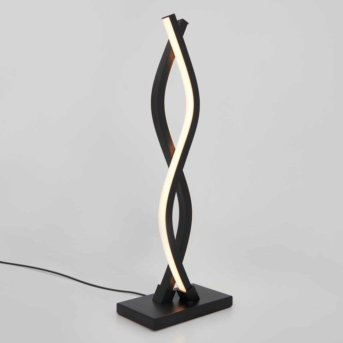 The Artika Wave LED Table Lamp showcases a modern, twisted ribbon design with a black base and frame and a spiraling white light strip. Plugged in, it’s displayed against a plain white background.