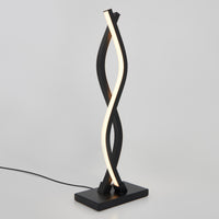 The Artika Wave LED Table Lamp showcases a modern, twisted ribbon design with a black base and frame and a spiraling white light strip. Plugged in, it’s displayed against a plain white background.
