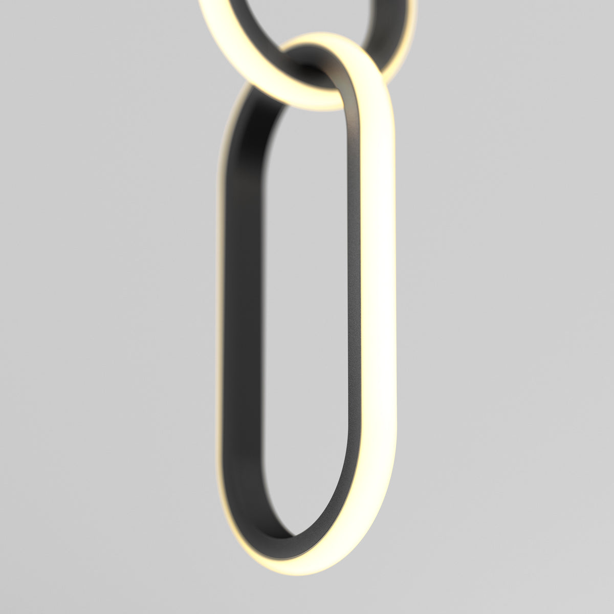 A close-up of Artikas Catena LED pendant light 5 CCT shows two interlocking, oval rings with a modern black finish, emitting a soft white edge glow, set against a plain light gray background.