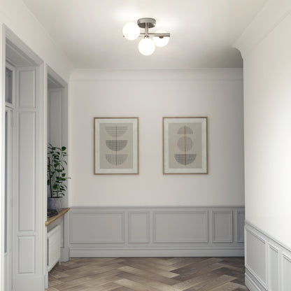 A hallway with light gray wainscoting, herringbone wood flooring, two abstract framed prints, and a potted plant on the window ledge is illuminated by the Bloom LED flush mount ceiling light from Artika.