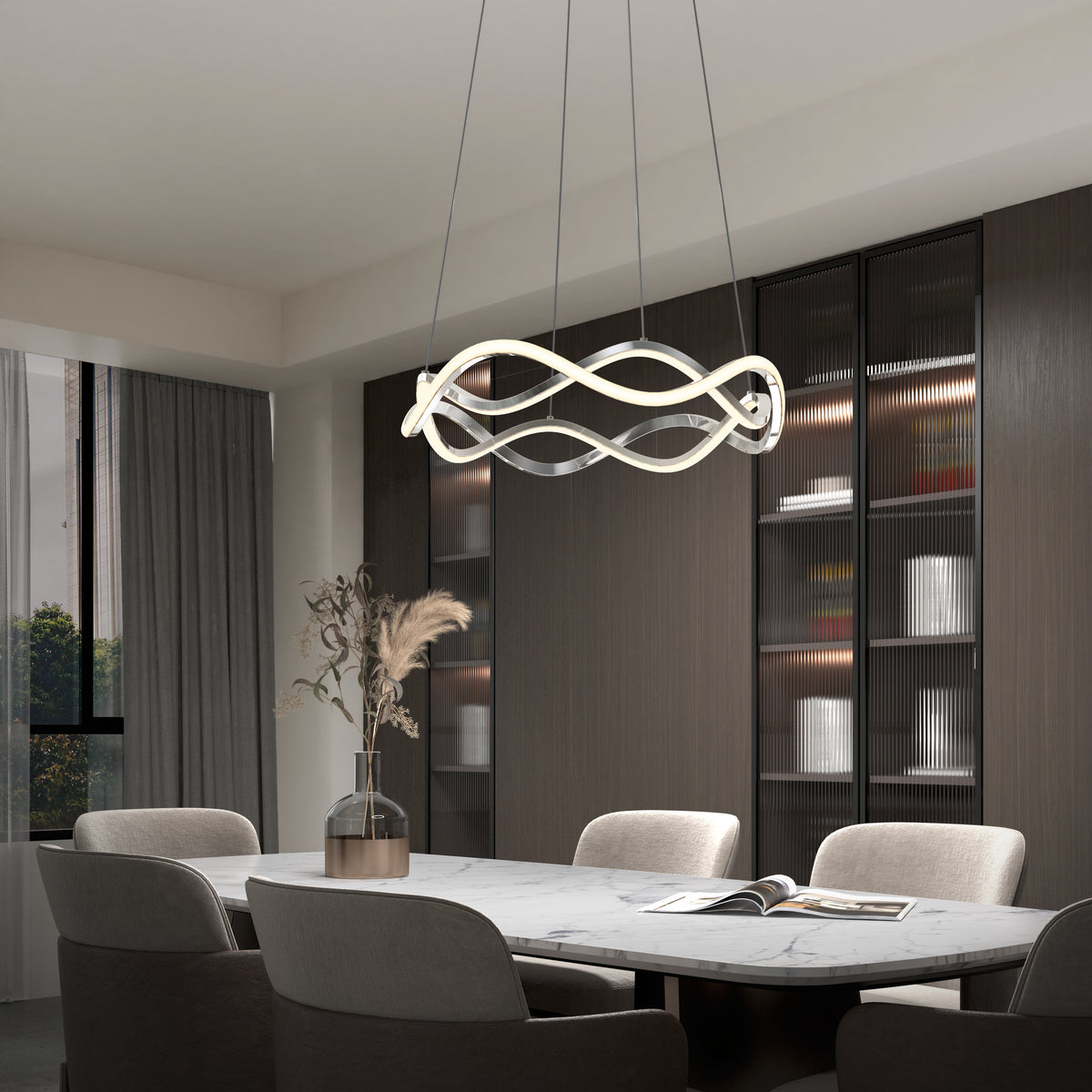 Modern dining room featuring an Artika Vanora LED pendant light with 5 CCT settings, a marble table, four beige chairs, a vase with dried flowers, and a bookshelf with glass doors. An open book rests on the table.