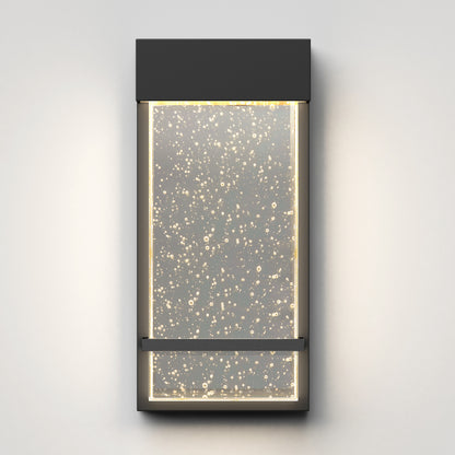 The Artika Skyler indoor-outdoor LED wall light features a modern rectangular black frame and a clear bubble panel, softly lit with warm LEDs and shown mounted on a light grey wall.
