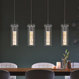 Four modern cylindrical pendant lights with textured glowing centers hang above a wooden dining table set with candles, a copper pitcher, and plants. The scene features gray upholstered chairs and a dark wall background.