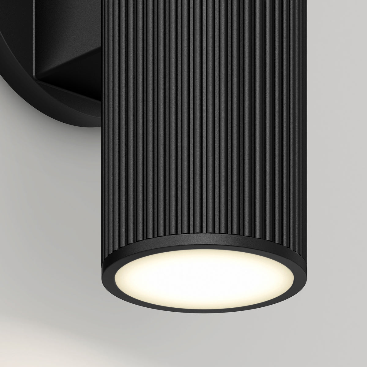 A close-up of a modern black cylindrical wall sconce with vertical grooves, mounted on a light gray wall, emitting a soft, warm white light from its circular base.